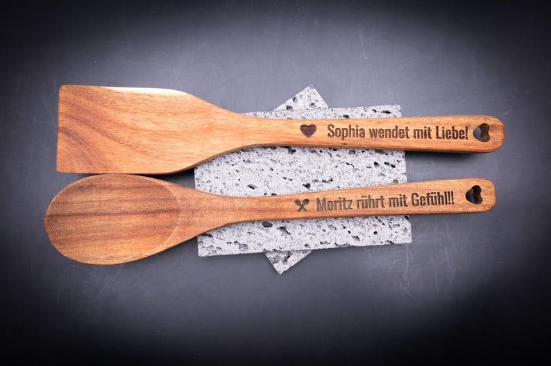 Flach gelegtes 2er-Set gravierter Kochlöffel aus Akazienholz mit Personalisierung. Gravuren „Sophia wendet mit Liebe!“ und „Moritz rührt mit Gefühl!!!“ mit Symbolen und Herzloch im Griff. Perfektes Geschenk für Kochfans, Paare oder Familien – nachhaltig, stilvoll und mit individueller Gravur.