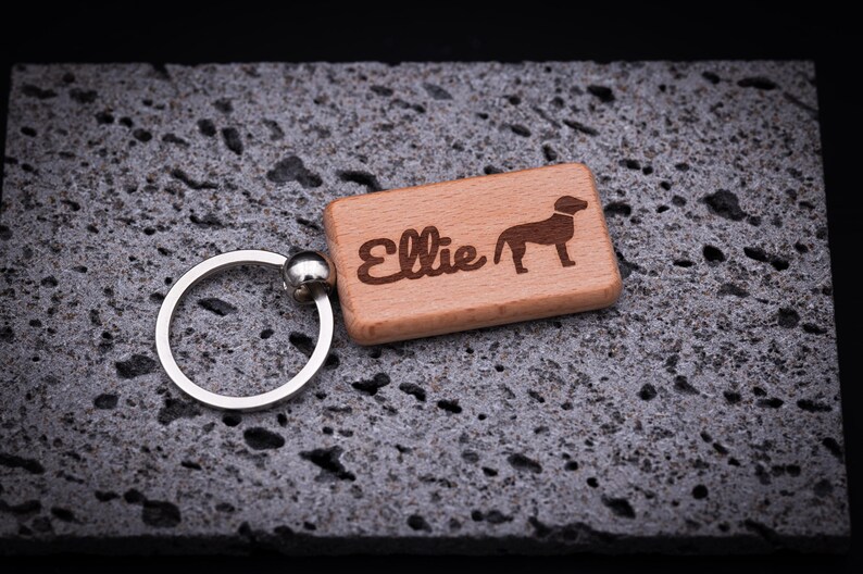 Rechteckiger Schlüsselanhänger aus massivem Buchenholz, graviert mit dem Namen Ellie und einem Hundesymbol. Der Schlüsselanhänger misst ca. 5 cm Länge, hat eine glatte Oberfläche, abgerundete Kanten und ist mit einem stabilen Schlüsselring aus Edelstahl ausgestattet. Eine individuelle Geschenkidee für Hundefreunde oder Tierliebhaber, perfekt als personalisierter Begleiter für Schlüssel, Tasche oder Rucksack mit langlebiger Lasergravur.