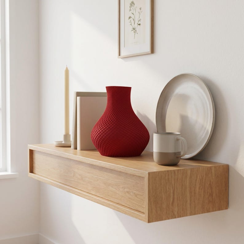 May include: A modern, floating wooden shelf with a red textured vase, a white plate, and a two-tone mug. A framed botanical print and a tall candle add to the minimalist decor. The shelf is mounted on a white wall.