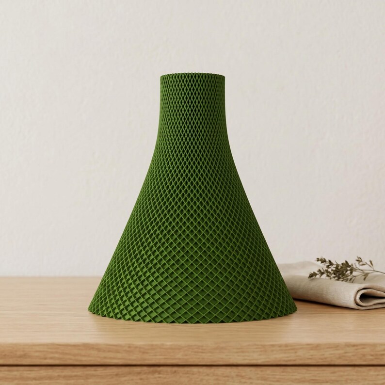 Peut inclure: Un vase vert en forme de c&ocirc;ne avec un motif en treillis. Le vase a une base large qui se r&eacute;tr&eacute;cit vers le haut. Le vase est fait d'un mat&eacute;riau vert uni et est pos&eacute; sur une surface en bois.