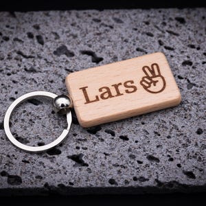 Rechteckiger Schlüsselanhänger aus naturbelassenem Buchenholz, ca. 5 cm lang, personalisiert mit dem Namen Lars und einem Peace-Symbol. Mit präziser Lasergravur gefertigt, stabil verarbeitet und mit abgerundeten Kanten für angenehme Haptik. Der hochwertige Schlüsselring aus Metall sorgt für sicheren Halt. Ein originelles Geschenk für Familie, Freunde oder als individuelles Accessoire für Schlüsselbund, Rucksack oder Handtasche.