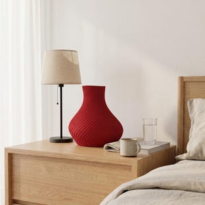 May include: A red decorative vase with a textured surface sits on a wooden nightstand. A beige lamp with a black base is to the left of the vase. A mug, a glass of water, and a book are also on the nightstand.