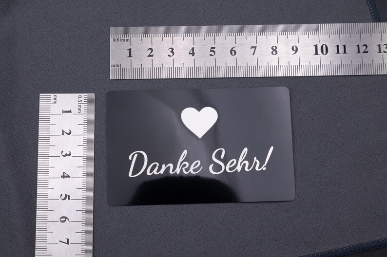 Schwarze Aluminiumkarte mit Gravur „Danke Sehr!“ und Herzsymbol, fotografiert mit Lineal zur Größenangabe. Format entspricht einer klassischen Kreditkarte (8,6 × 5,4 cm). Langlebig, robust und stilvoll – ideal als Dankeskarte oder personalisierte Visitenkarte für professionelle Businessauftritte.