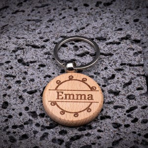 Runder Schlüsselanhänger aus Holz mit Gravur „Emma“. Der Name ist in einem zarten Rahmen mit Schnörkel-Ornamenten eingefasst. Natürliches Holzdesign mit stabilem Schlüsselring.
