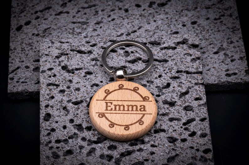 Runder Schlüsselanhänger aus Holz mit Gravur „Emma“. Der Name ist in einem zarten Rahmen mit Schnörkel-Ornamenten eingefasst. Natürliches Holzdesign mit stabilem Schlüsselring.
