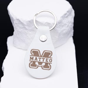 Personalisierter Schlüsselanhänger aus weißem veganem Kunstleder mit eleganter Lasergravur des Namens „Matteo“ im klassischen Monogramm-Stil. Das tropfenförmige Design mit silbernem Metallring und stabiler Niete verbindet Stil und Funktionalität. Das Foto zeigt den Anhänger auf heller Steinunterlage mit Lineal zur Größenangabe – ideal als hochwertiges, personalisierbares Accessoire oder besonderes Geschenk für Mann, Frau oder Kind.