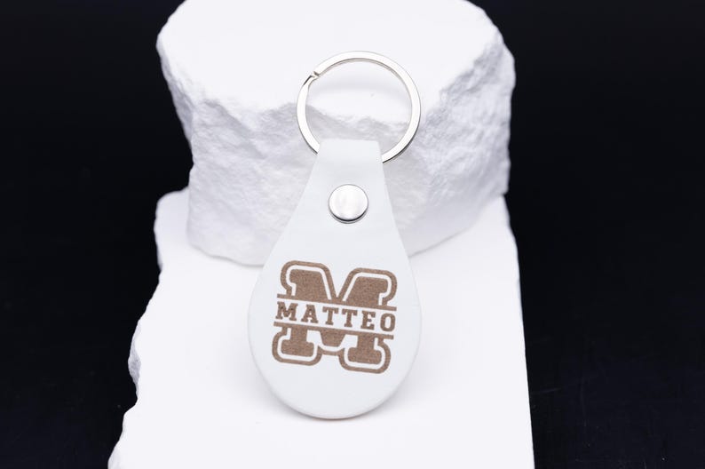 Personalisierter Schlüsselanhänger aus weißem veganem Kunstleder mit eleganter Lasergravur des Namens „Matteo“ im klassischen Monogramm-Stil. Das tropfenförmige Design mit silbernem Metallring und stabiler Niete verbindet Stil und Funktionalität. Das Foto zeigt den Anhänger auf heller Steinunterlage mit Lineal zur Größenangabe – ideal als hochwertiges, personalisierbares Accessoire oder besonderes Geschenk für Mann, Frau oder Kind.