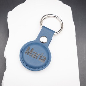 Blauer AirTag Schlüsselanhänger aus PU Leder mit individueller Lasergravur des Namens Maria. Die Gravur ist kontrastreich und dauerhaft eingebrannt. Ausgestattet mit stabilem Edelstahl Schlüsselring und Druckknopf. Moderner personalisierter Schlüsselanhänger für Apple AirTag, ideal als Geschenk für Frauen Männer oder Kinder.