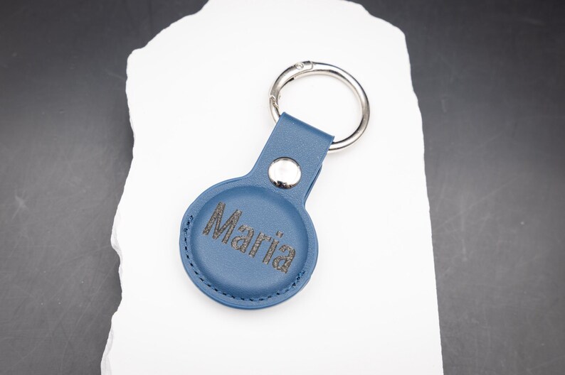 Blauer AirTag Schlüsselanhänger aus PU Leder mit individueller Lasergravur des Namens Maria. Die Gravur ist kontrastreich und dauerhaft eingebrannt. Ausgestattet mit stabilem Edelstahl Schlüsselring und Druckknopf. Moderner personalisierter Schlüsselanhänger für Apple AirTag, ideal als Geschenk für Frauen Männer oder Kinder.
