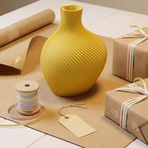 Puede incluir: Un jarr&oacute;n amarillo con un dise&ntilde;o en espiral se encuentra sobre una superficie marr&oacute;n, junto a una bobina de cinta de colores pastel y dos regalos envueltos. Tambi&eacute;n hay una etiqueta en blanco y un rollo de papel marr&oacute;n.