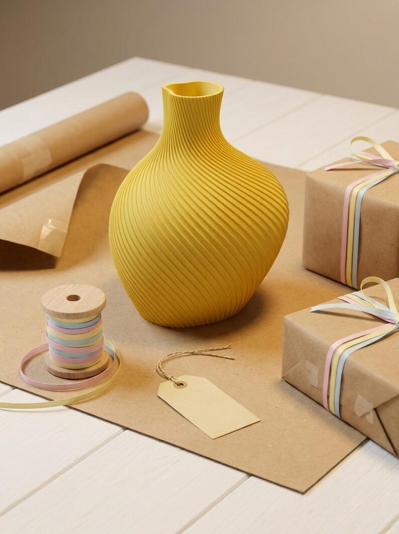 Puede incluir: Un jarr&oacute;n amarillo con un dise&ntilde;o en espiral se encuentra sobre una superficie marr&oacute;n, junto a una bobina de cinta de colores pastel y dos regalos envueltos. Tambi&eacute;n hay una etiqueta en blanco y un rollo de papel marr&oacute;n.