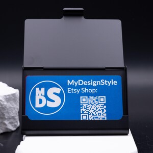 Geöffnetes schwarzes Metall-Visitenkartenetui mit einer blauen Businesskarte im Inneren. Karte zeigt Logo, QR-Code und Etsy-Shop. Das Etui kombiniert moderne Eleganz mit praktischer Funktion, ist leicht und robust. Perfekt für Geschäftsleute, die Karten professionell präsentieren möchten. Individuell gravierbar, ideal als Geschenk oder stilvolles Business-Accessoire für Büro und Meetings.