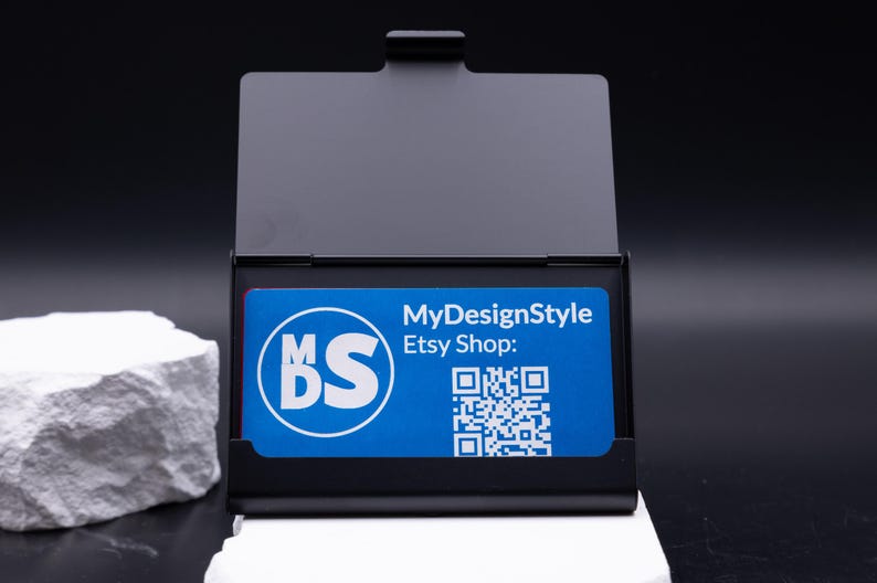 Geöffnetes schwarzes Metall-Visitenkartenetui mit einer blauen Businesskarte im Inneren. Karte zeigt Logo, QR-Code und Etsy-Shop. Das Etui kombiniert moderne Eleganz mit praktischer Funktion, ist leicht und robust. Perfekt für Geschäftsleute, die Karten professionell präsentieren möchten. Individuell gravierbar, ideal als Geschenk oder stilvolles Business-Accessoire für Büro und Meetings.