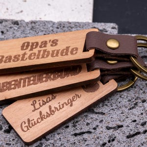 Drei personalisierte Schlüsselanhänger aus Holz mit braunem Kunstlederband und Messingring, übereinander gestapelt auf grauer Steinplatte. Gravierte Aufschriften: „Opa’s Bastelbude“, „Abenteuerbund“ und „Lisas Glücksbringer“. Die natürliche Holzoptik kombiniert mit den dunklen Lederriemen und goldfarbenen Metallringen macht die Anhänger robust, dekorativ und zu idealen Geschenkideen für Familie oder Freunde.