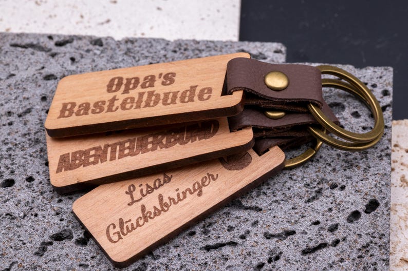 Drei personalisierte Schlüsselanhänger aus Holz mit braunem Kunstlederband und Messingring, übereinander gestapelt auf grauer Steinplatte. Gravierte Aufschriften: „Opa’s Bastelbude“, „Abenteuerbund“ und „Lisas Glücksbringer“. Die natürliche Holzoptik kombiniert mit den dunklen Lederriemen und goldfarbenen Metallringen macht die Anhänger robust, dekorativ und zu idealen Geschenkideen für Familie oder Freunde.