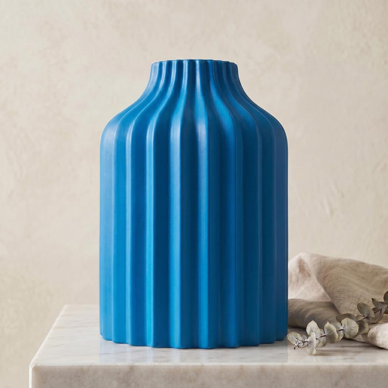 Może przedstawiać: Żywy niebieski ceramiczny wazon z żebrowanym wzorem. Wazon ma cylindryczny kształt z lekko rozszerzonym wierzchołkiem. Stoi na marmurowej powierzchni, z neutralną kolorystycznie tkaniną i gałązką zieleni w tle.