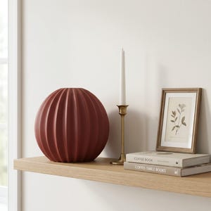 May include: A terracotta-coloured, ribbed spherical vase sits on a wooden shelf, next to a gold candlestick with a white candle. A framed botanical print and two books complete the display. Home decor.