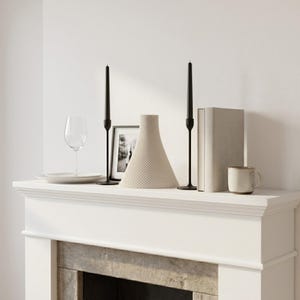 Peut inclure: Une composition d&eacute;corative sur une chemin&eacute;e blanche. Elle comprend un vase beige textur&eacute;, deux bougeoirs noirs avec des bougies noires, une photo encadr&eacute;e, un verre &agrave; vin, une assiette, un livre et une tasse.