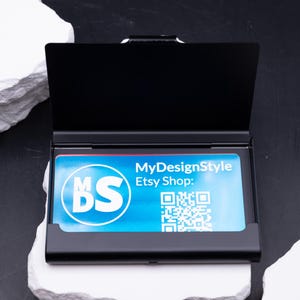 Schwarzes Metall-Visitenkartenetui halb geöffnet, eine blaue Businesskarte ist sichtbar. Vorderseite zeigt Lasergravur „meinefirma.de“. Robustes, leichtes Etui, das Karten zuverlässig schützt. Individuell gravierbar mit Logo, Initialen oder Firmenname. Perfektes Geschenk für Geschäftspartner, Kollegen oder Kunden. Praktisch für Büro, Networking und Business-Alltag.