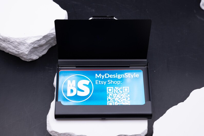 Schwarzes Metall-Visitenkartenetui halb geöffnet, eine blaue Businesskarte ist sichtbar. Vorderseite zeigt Lasergravur „meinefirma.de“. Robustes, leichtes Etui, das Karten zuverlässig schützt. Individuell gravierbar mit Logo, Initialen oder Firmenname. Perfektes Geschenk für Geschäftspartner, Kollegen oder Kunden. Praktisch für Büro, Networking und Business-Alltag.