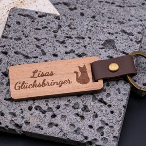 Schlüsselanhänger aus Holz mit Gravur „Lisas Glücksbringer“ und Katzen-Symbol. Der rechteckige Holzanhänger ist mit braunem Kunstlederband und stabilem Messingring versehen und kombiniert natürliche Holzoptik mit edlem Lederlook. Durch die individuelle Gravur wird er zum persönlichen Geschenk für Katzenliebhaber, Freunde oder Familie. Praktisches Accessoire, langlebig und ideal für Schlüsselbund oder Alltag.