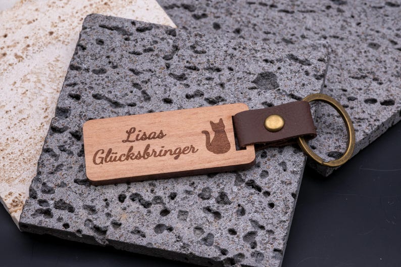 Schlüsselanhänger aus Holz mit Gravur „Lisas Glücksbringer“ und Katzen-Symbol. Der rechteckige Holzanhänger ist mit braunem Kunstlederband und stabilem Messingring versehen und kombiniert natürliche Holzoptik mit edlem Lederlook. Durch die individuelle Gravur wird er zum persönlichen Geschenk für Katzenliebhaber, Freunde oder Familie. Praktisches Accessoire, langlebig und ideal für Schlüsselbund oder Alltag.
