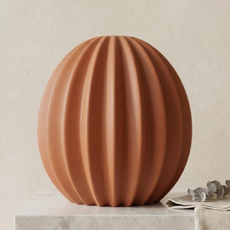 May include: A terracotta-coloured, spherical vase with vertical, ribbed detailing. The vase has a rounded shape and sits on a marble surface. The neutral colour and textured design make it a versatile decorative piece.