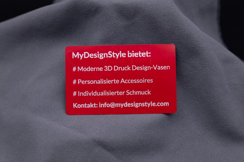 Rote Aluminiumkarte mit Gravur von Leistungen: moderne 3D-Druck-Designvasen, personalisierte Accessoires, individueller Schmuck. Darunter die Kontaktadresse von MyDesignStyle. Diese Metallkarte ist eine exklusive und langlebige Möglichkeit, das eigene Produktportfolio und die Marke stilvoll zu präsentieren.
