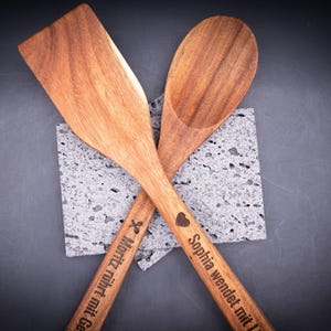 Kreuzweise angeordnete Kochlöffel aus Akazienholz mit personalisierter Gravur – einer mit Herzsymbol und Spruch „Sophia wendet mit Liebe!“, der andere mit Symbol und Spruch „Moritz rührt mit Gefühl!!!“. Stilvolles Küchenaccessoire aus natürlichem Holz, individuell gravierbar mit Namen oder Botschaft.
