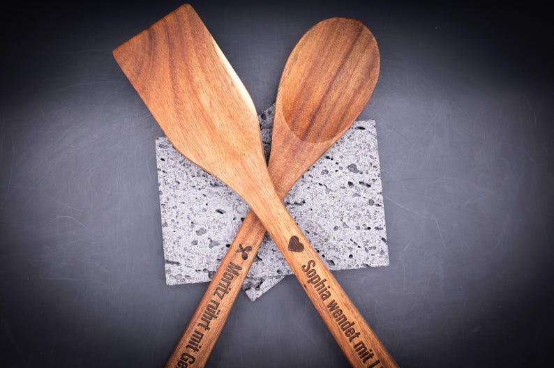 Kreuzweise angeordnete Kochlöffel aus Akazienholz mit personalisierter Gravur – einer mit Herzsymbol und Spruch „Sophia wendet mit Liebe!“, der andere mit Symbol und Spruch „Moritz rührt mit Gefühl!!!“. Stilvolles Küchenaccessoire aus natürlichem Holz, individuell gravierbar mit Namen oder Botschaft.