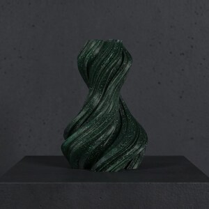 K&ouml;nnte beinhalten: Eine dunkelgr&uuml;ne, abstrakte Vase mit einem wirbelnden, strukturierten Design. Die Vase hat ein gesprenkeltes Aussehen mit kleinen wei&szlig;en Flecken. Sie wird auf einer dunkelgrauen Oberfl&auml;che vor einem passenden Hintergrund pr&auml;sentiert. Die Vase hat eine einzigartige, k&uuml;nstlerische Form.