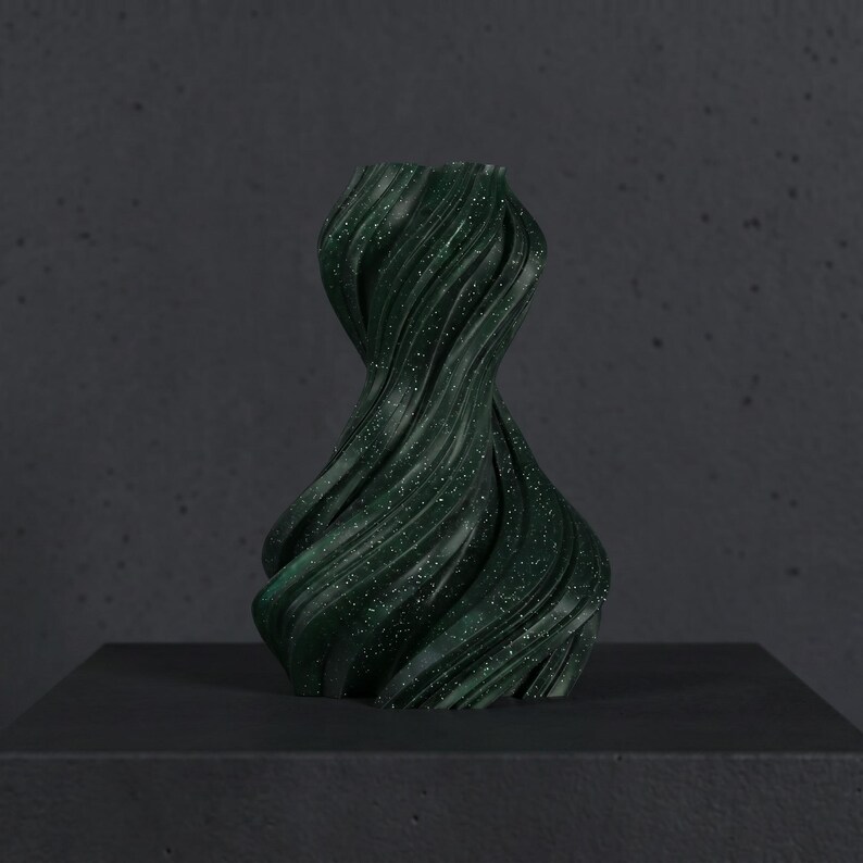 K&ouml;nnte beinhalten: Eine dunkelgr&uuml;ne, abstrakte Vase mit einem wirbelnden, strukturierten Design. Die Vase hat ein gesprenkeltes Aussehen mit kleinen wei&szlig;en Flecken. Sie wird auf einer dunkelgrauen Oberfl&auml;che vor einem passenden Hintergrund pr&auml;sentiert. Die Vase hat eine einzigartige, k&uuml;nstlerische Form.