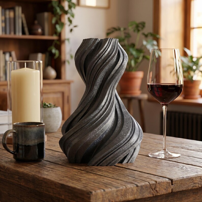 K&ouml;nnte beinhalten: Eine dunkelgraue, strukturierte Vase mit einem Wirbeldesign steht auf einem Holztisch. Ein Glas Rotwein, eine Tasse und eine hohe Kerze in einem Glashalter stehen ebenfalls auf dem Tisch. Der Hintergrund umfasst ein B&uuml;cherregal und Pflanzen.