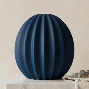 May include: A navy blue, spherical vase with vertical, ribbed detailing. The vase is made of a matte material and sits on a white marble surface. The vase has a rounded shape and is a decorative home accent.