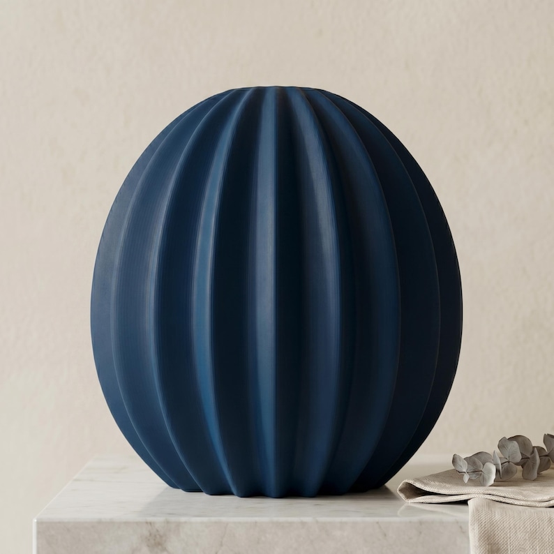May include: A navy blue, spherical vase with vertical, ribbed detailing. The vase is made of a matte material and sits on a white marble surface. The vase has a rounded shape and is a decorative home accent.