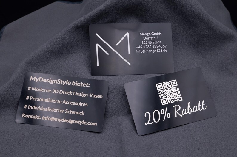Drei exklusive Visitenkarten aus schwarzem Aluminium mit Lasergravur. Links gravierte Infos zu angebotenen Produkten, in der Mitte eine Businesskarte mit Logo und Kontaktdaten, rechts eine Rabattkarte mit QR-Code und Gravur „20% Rabatt“. Die hochwertigen Metallkarten sind stabil, elegant und modern – ideal als personalisierte Businesskarten, Werbekarten oder Kundenkarten.