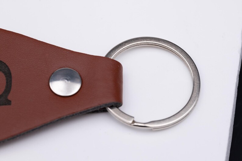 Nahaufnahme des oberen Bereichs des personalisierten Schlüsselanhängers mit Fokus auf den silberfarbenen Metallring und die robuste Niete. Das braune Kunstleder zeigt feine Struktur und präzise Schnittkanten. Diese Detailaufnahme verdeutlicht die hochwertige Verarbeitung und Langlebigkeit des Designs – ideal für den täglichen Gebrauch und als Geschenk für Menschen, die Qualität und Stil schätzen.