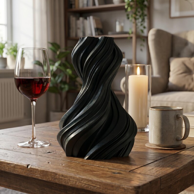 K&ouml;nnte beinhalten: Eine schwarze, skulpturale Vase mit einem gedrehten Design steht auf einem Holztisch. Ein Glas Rotwein und eine brennende Kerze in einem Glashalter stehen ebenfalls auf dem Tisch. Rechts von der Vase befindet sich eine Tasse.