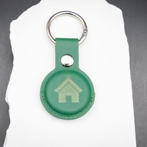 Personalisierter AirTag Schlüsselanhänger aus grünem PU Leder mit graviertem Haus Symbol auf der Vorderseite. Der Schlüsselanhänger liegt auf einem weißen Stein und ist mit einem stabilen Edelstahl Schlüsselring ausgestattet. Die runde Form passt exakt für Apple AirTag. Ideal zur Kennzeichnung von Hausschlüsseln oder als praktisches Geschenk für Familie oder Freunde.
