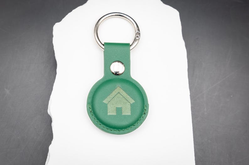 Personalisierter AirTag Schlüsselanhänger aus grünem PU Leder mit graviertem Haus Symbol auf der Vorderseite. Der Schlüsselanhänger liegt auf einem weißen Stein und ist mit einem stabilen Edelstahl Schlüsselring ausgestattet. Die runde Form passt exakt für Apple AirTag. Ideal zur Kennzeichnung von Hausschlüsseln oder als praktisches Geschenk für Familie oder Freunde.
