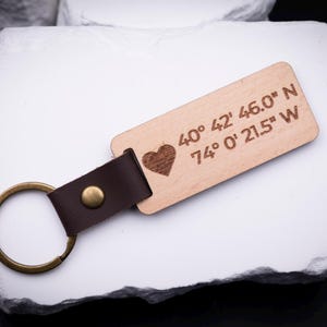 Ein gravierter Schlüsselanhänger aus hellem Holz mit Herzsymbol und Koordinaten „40° 42' 46.0'' N, 74° 0' 21.5'' W“ liegt auf weißem Stein. Der Anhänger ist durch ein braunes Lederband mit goldfarbenem Metallring verbunden. Ideal für individuelle Gravuren mit Ortsangaben, Namen oder Botschaften. Ein stilvolles, handgefertigtes Accessoire, das persönliche Erinnerungen festhält und sich perfekt als Geschenk eignet.