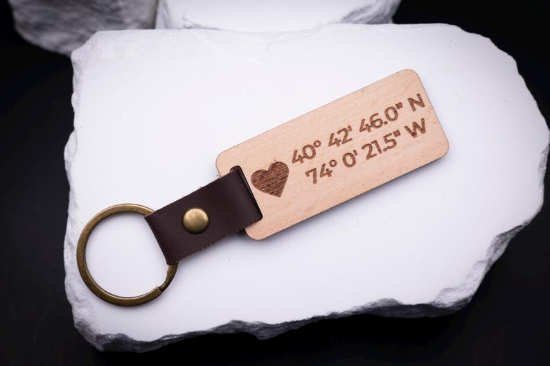 Ein gravierter Schlüsselanhänger aus hellem Holz mit Herzsymbol und Koordinaten „40° 42' 46.0'' N, 74° 0' 21.5'' W“ liegt auf weißem Stein. Der Anhänger ist durch ein braunes Lederband mit goldfarbenem Metallring verbunden. Ideal für individuelle Gravuren mit Ortsangaben, Namen oder Botschaften. Ein stilvolles, handgefertigtes Accessoire, das persönliche Erinnerungen festhält und sich perfekt als Geschenk eignet.
