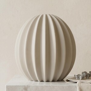 May include: A beige, spherical decorative vase with vertical, ribbed detailing. The vase is displayed on a marble surface, with a neutral background. The vase has a modern, minimalist design, suitable for home decor.