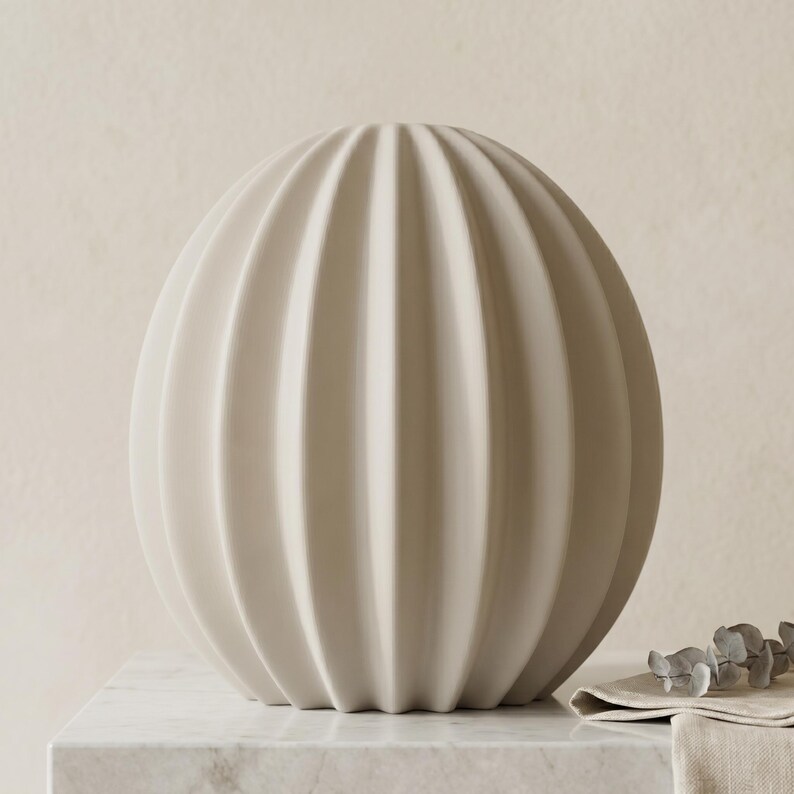 May include: A beige, spherical decorative vase with vertical, ribbed detailing. The vase is displayed on a marble surface, with a neutral background. The vase has a modern, minimalist design, suitable for home decor.