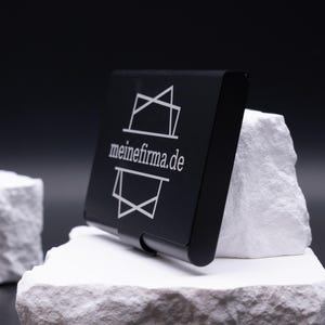 Schwarzes Metall-Visitenkartenetui schräg auf weißem Stein, Vorderseite mit Lasergravur „meinefirma.de“. Hochwertig verarbeitet, elegant und leicht. Ideal für die Personalisierung mit Logo, Initialen oder Firmennamen. Ein stilvolles Business-Accessoire für Unternehmer, Kollegen und Geschäftspartner. Perfekt als Geschenkidee für Männer und Frauen im Büro- oder Businessalltag.