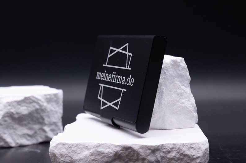 Schwarzes Metall-Visitenkartenetui schräg auf weißem Stein, Vorderseite mit Lasergravur „meinefirma.de“. Hochwertig verarbeitet, elegant und leicht. Ideal für die Personalisierung mit Logo, Initialen oder Firmennamen. Ein stilvolles Business-Accessoire für Unternehmer, Kollegen und Geschäftspartner. Perfekt als Geschenkidee für Männer und Frauen im Büro- oder Businessalltag.