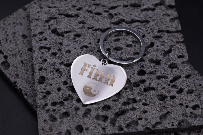 Glänzender Herz-Schlüsselanhänger aus Edelstahl mit Gravur „Finn“ und Yin-Yang-Motiv. Der Anhänger zeigt feine Lasergravurdetails und liegt auf strukturiertem, dunklem Stein. Ideal als symbolisches Geschenk für Ausgeglichenheit und Freundschaft – robust, langlebig und individuell personalisierbar.
