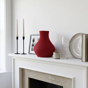 May include: A red textured vase, two black candle holders with beige candles, a framed picture, a small mug, a wine glass, a plate, and a book are arranged on a white mantelpiece. The vase has a unique woven pattern.