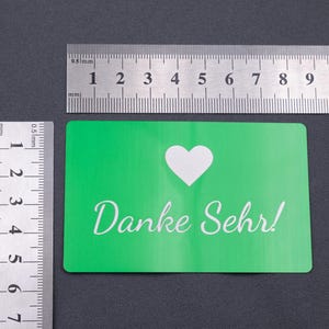 Grüne Metall-Visitenkarte mit „Danke Sehr!“ Gravur, daneben Lineale zur Größenanzeige. Die Karte entspricht klassischen Visitenkartenmaßen (ca. 8,5 x 5,4 cm) und ist aus stabilem Aluminium gefertigt. Das moderne, minimalistische Design kombiniert mit der widerstandsfähigen Oberfläche macht die Karte zu einer idealen Wahl für exklusive Geschäftsausstattung oder besondere Dankeskarten.