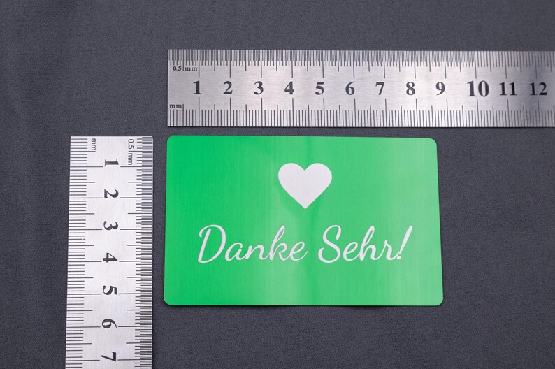 Grüne Metall-Visitenkarte mit „Danke Sehr!“ Gravur, daneben Lineale zur Größenanzeige. Die Karte entspricht klassischen Visitenkartenmaßen (ca. 8,5 x 5,4 cm) und ist aus stabilem Aluminium gefertigt. Das moderne, minimalistische Design kombiniert mit der widerstandsfähigen Oberfläche macht die Karte zu einer idealen Wahl für exklusive Geschäftsausstattung oder besondere Dankeskarten.
